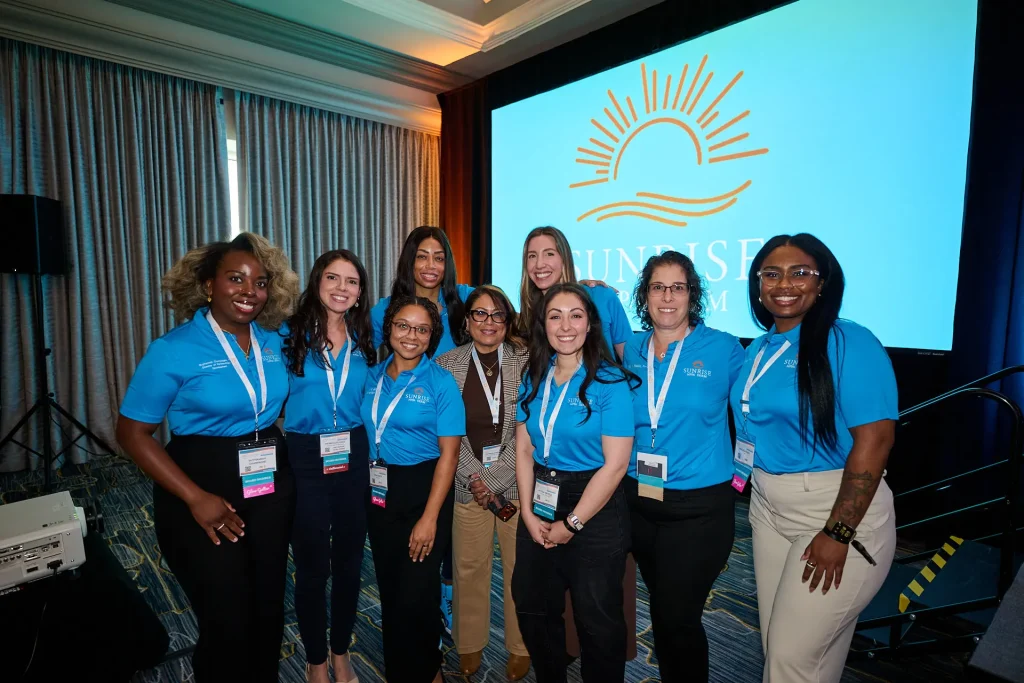 Eight women posing together in front of a Sunrise NPPA Derm screen, most wearing matching blue branded shirts.