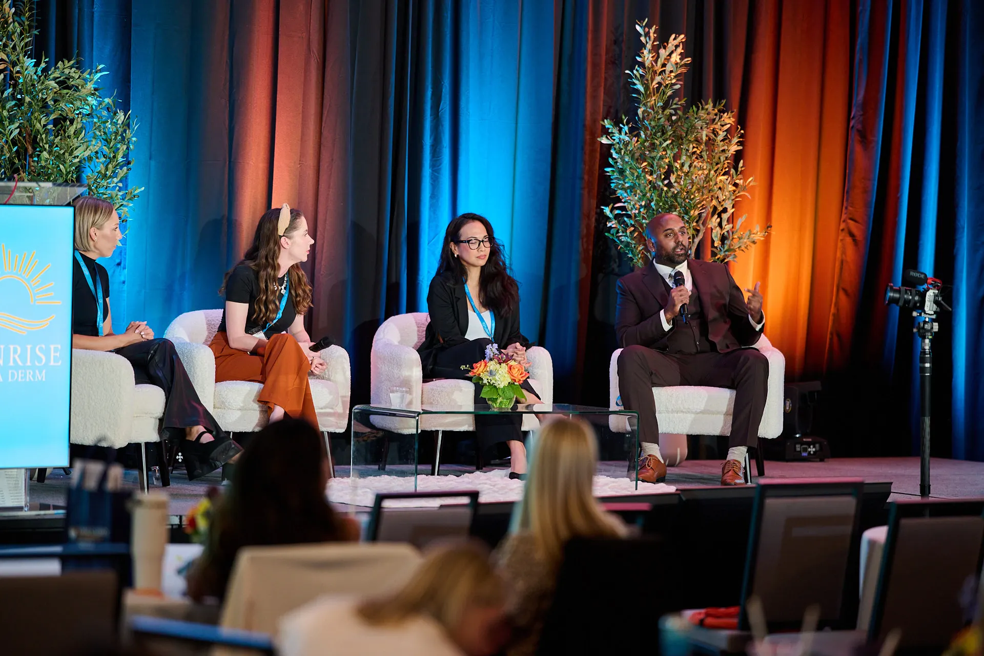Four panelists seated onstage at a Sunrise NPPA Derm conference, with one speaking into a microphone.